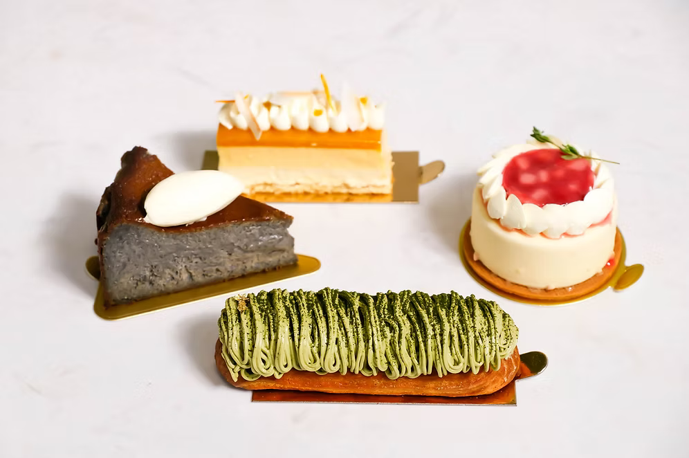 Assorted desserts including a green tea cake, a slice of chocolate cake, and a small white cake with strawberry on a white background.