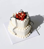 A two-tier white frosted cake decorated with fresh strawberries on top and a sheer black ribbon, displayed on a white square board against a white background. by Blue Moon Christchurch