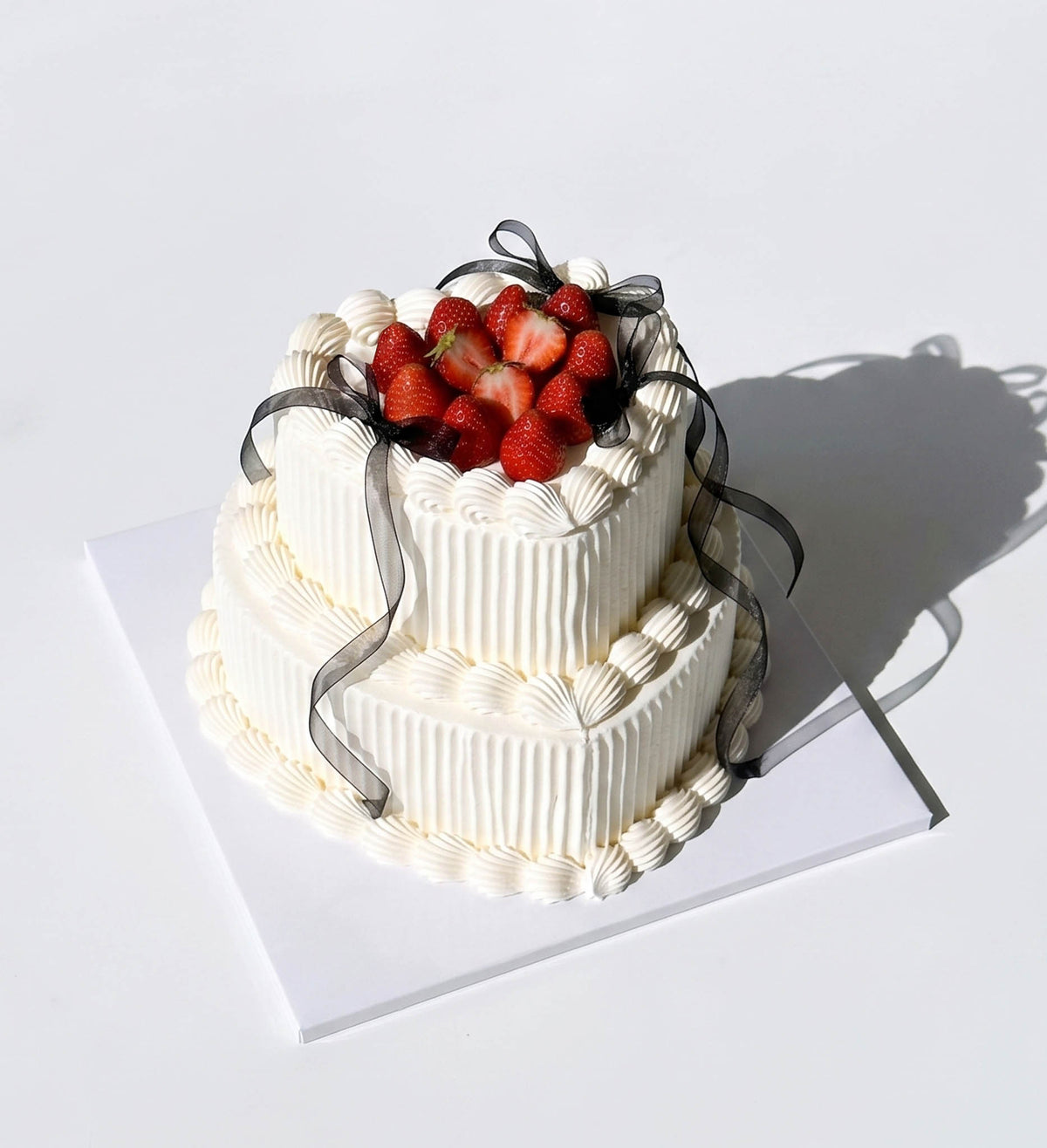A two-tier white frosted cake decorated with fresh strawberries on top and a sheer black ribbon, displayed on a white square board against a white background. by Blue Moon Christchurch
